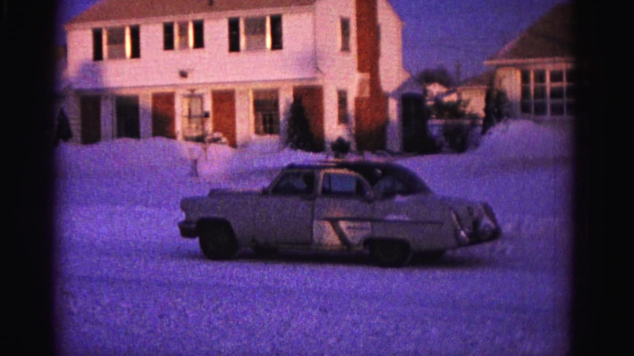 Tan sedan in snowy suburban driveway, white house with lit upper windows, pronounced purple color cast on 8mm film.