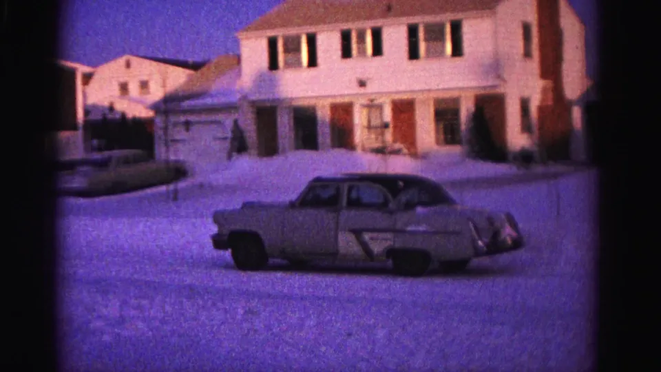 Early 1960s 8mm home movie — wider suburban street view, tan sedan prominent in foreground, multiple postwar houses in background, Hagerstown MD area, 1962.