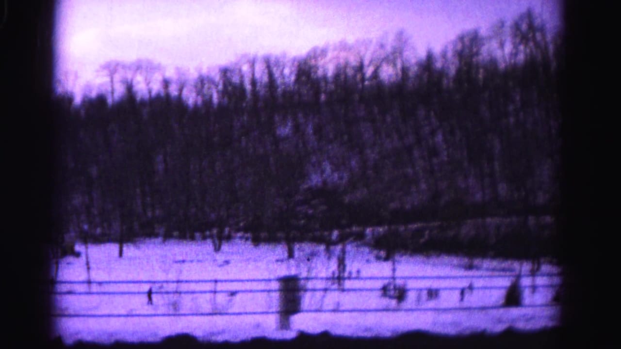 Underexposed 8mm home movie frame from approximately 1962 showing a snow-covered hillside, bare trees, and a fence line with a pronounced purple dye-shift.