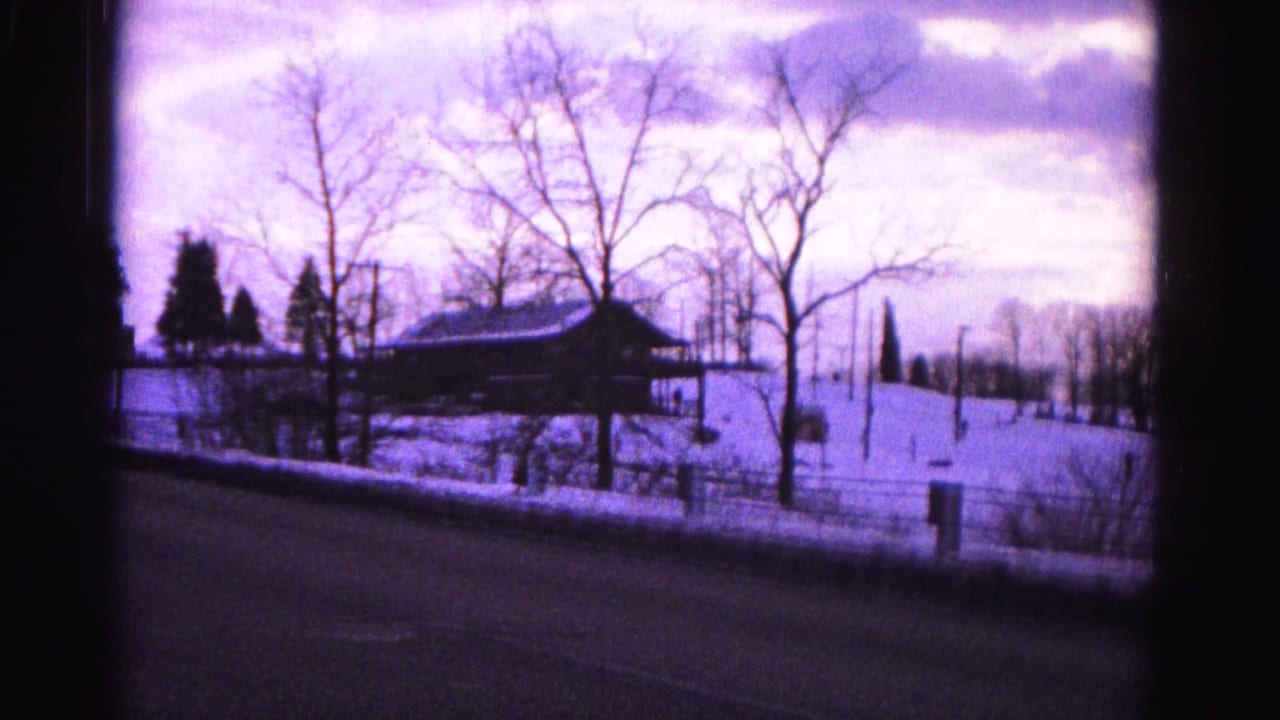 Rural road in winter with snow on the ground, a farm pavilion structure in the middle distance, bare trees, and a fence line; purple-tinted 8mm film.