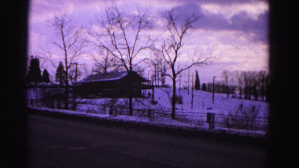 8mm home movie frame circa 1962 of a snow-covered rural landscape with a farm pavilion structure, bare trees, and a road edge; strong purple dye-shift cast.
