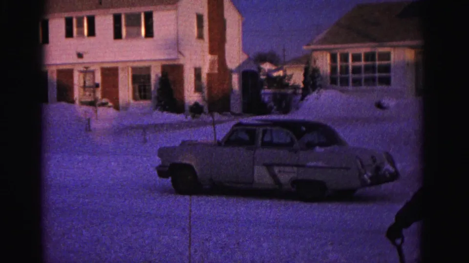 8mm home movie frame circa 1962 showing a two-story home with illuminated windows, a period American sedan in the snowy driveway, and figures at the right frame edge.