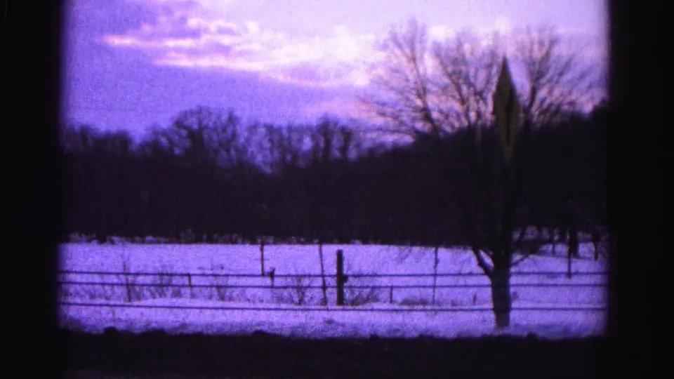 8mm home-movie frame circa 1962 showing a snow-covered field, wire fence, a bare deciduous tree, and a diamond-shaped road sign on a post; Hagerstown, Maryland area per metadata.