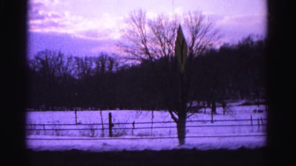 8mm home-movie frame circa 1962 with a diamond-shaped road sign and bare tree prominent beside a snow-covered field and wire fence; Hagerstown, Maryland area per metadata.