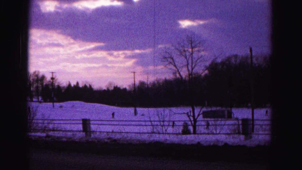 Snow-covered slope at dusk seen through a dark window frame, with barbed wire fence, bare trees, distant silhouetted figures on the hill, and a purple sky.