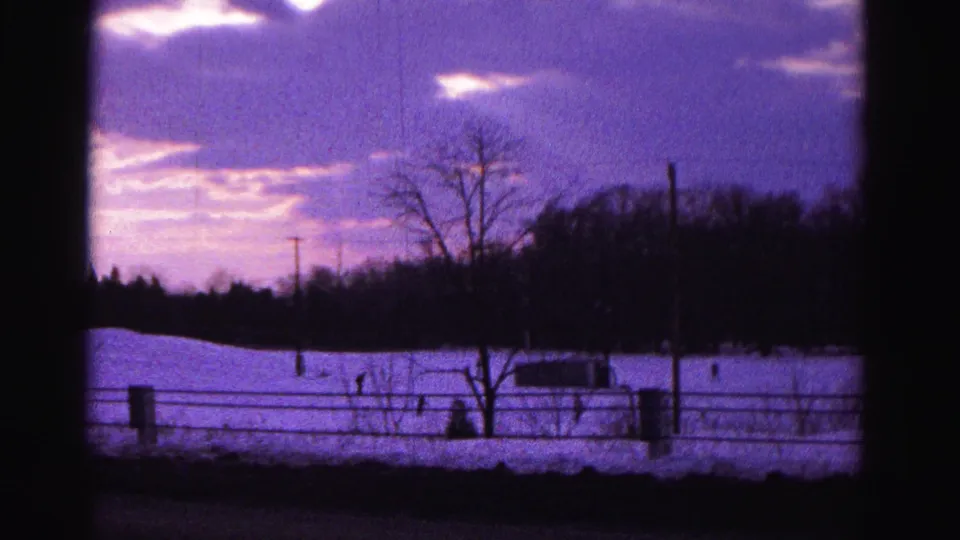8mm home movie frame from approximately 1962 showing a snow-covered rural slope at dusk near Hagerstown, Maryland, with distant silhouetted figures and a violet-cast sky.