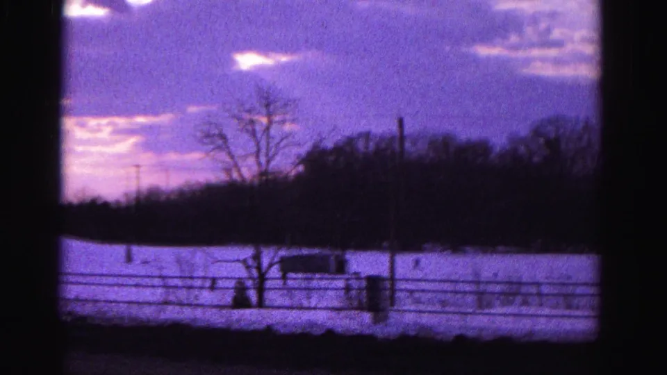 8mm home movie frame circa 1962 capturing a snow-covered field at dusk with a prominent fence line, dark structure, and a figure near the foreground, Hagerstown area.