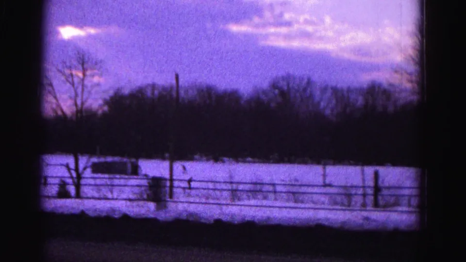 8mm home movie frame from approximately 1962 showing a snow-covered rural slope at dusk with a small figure visible on the right, fence, and treeline, Hagerstown area.