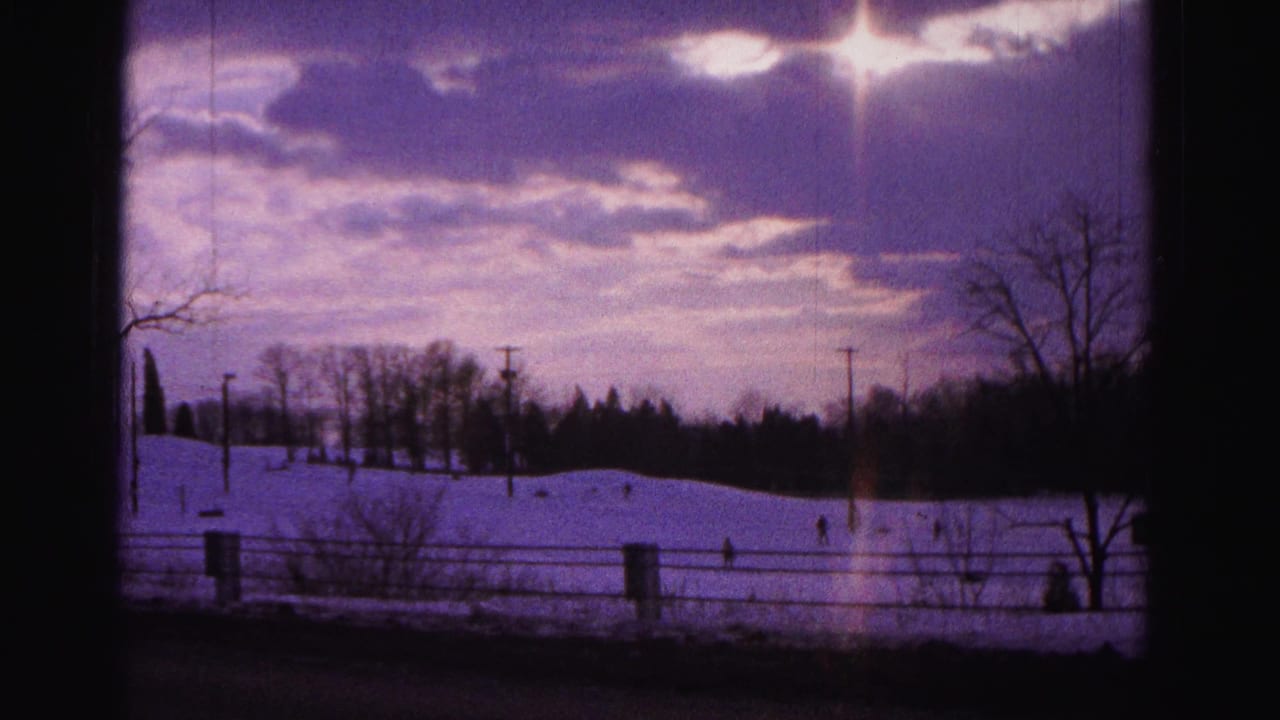 1962 8mm home movie frame showing a snow-covered hillside near Hagerstown, Maryland, with silhouetted figures on the slope, purple winter sky, and lens flare from a low sun.