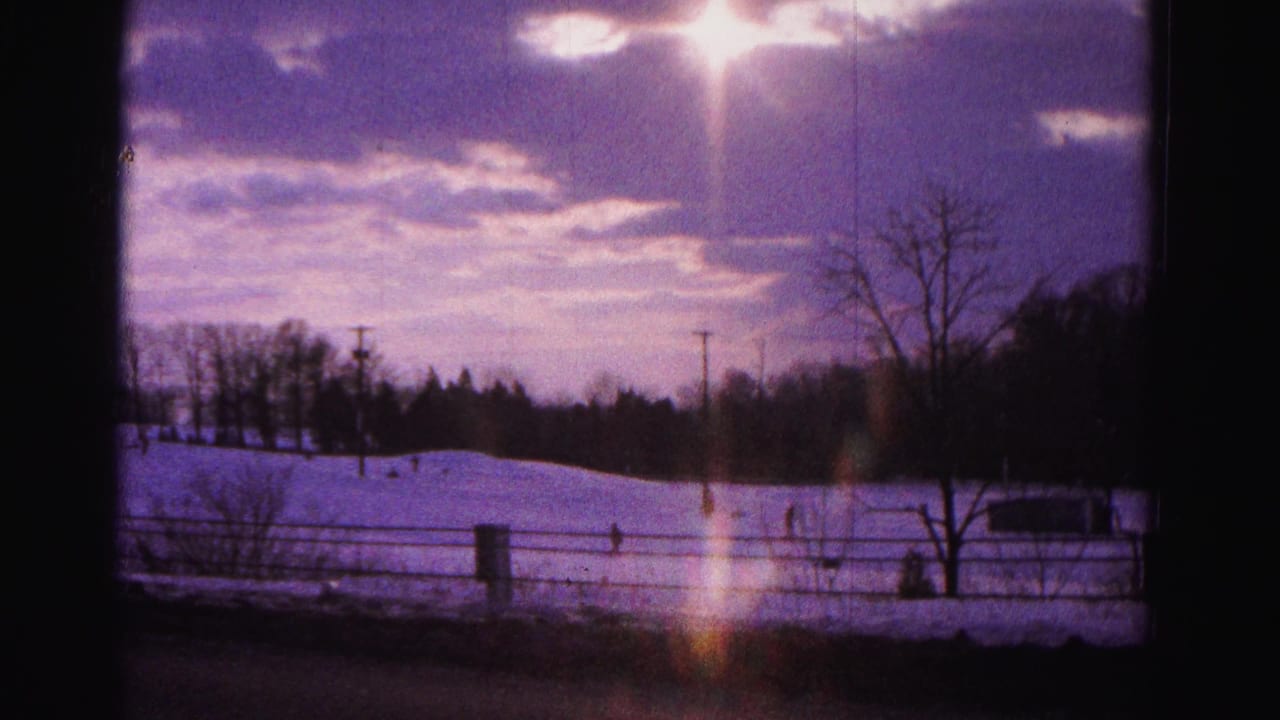 Wide snow-covered field through window frame, strong orange sun lens flare at right, purple-toned sky, bare trees and fence silhouetted, small distant figures.