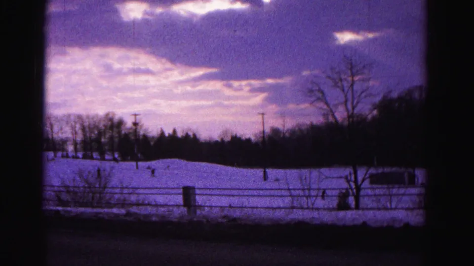 1962 8mm film frame capturing a snowy hillside near Hagerstown, Maryland, under a vivid purple winter sky, with bare trees, a fence line, and small silhouetted figures on the slope.