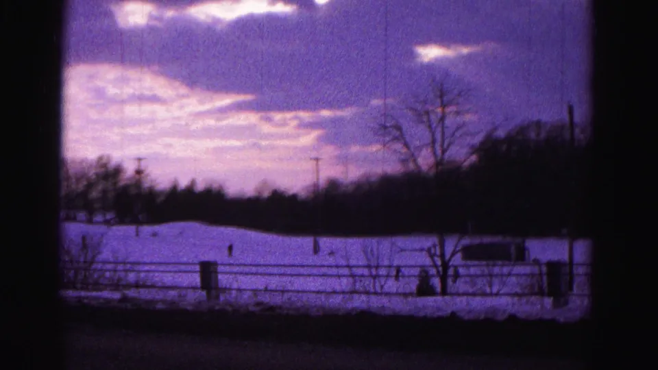 Final frame of 1962 8mm clip near Hagerstown, Maryland: snow-covered hillside under a purple dusk sky, with fence line, bare trees, utility poles, and distant silhouetted figures.