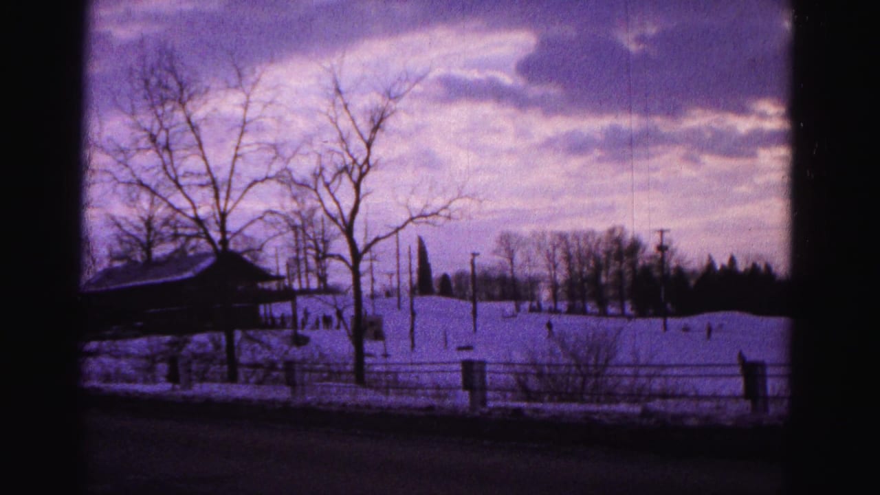 Snow-covered hill with distant figures, bare trees, wooden fence, roofed shelter, and purple-tinted cloudy sky — 8mm frame.