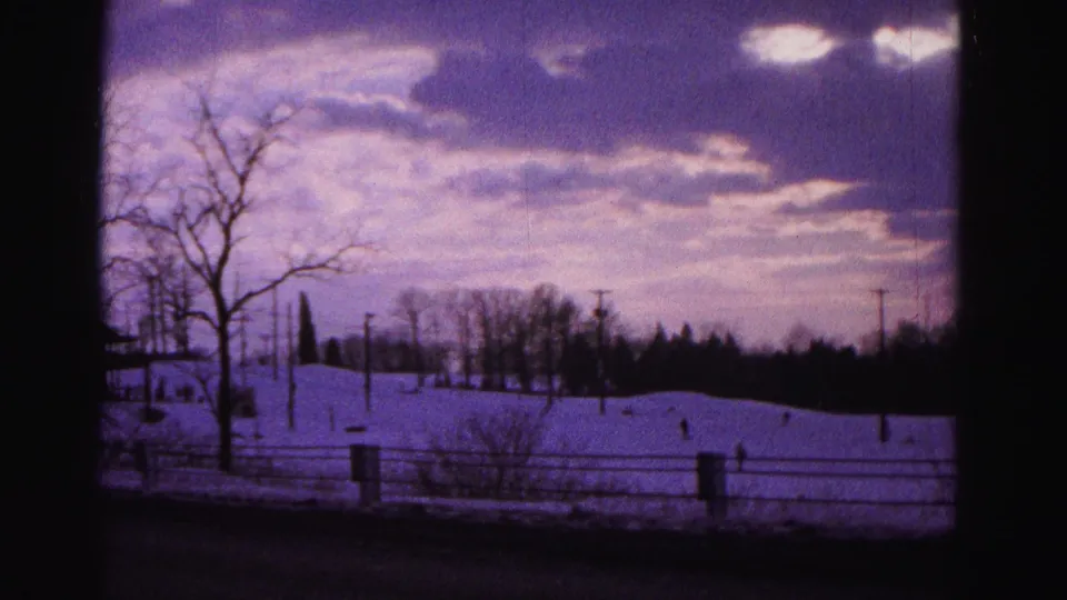 8mm home movie frame, 1962 Hagerstown Maryland — static wide shot of snowy slope and treeline with wooden fence, utility poles, and aged purple sky.