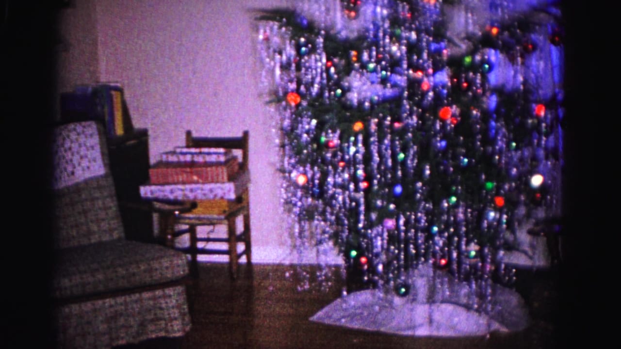 1962 Hagerstown, Maryland home-movie frame showing an aluminum Christmas tree with colorful ornaments and tinsel beside an upholstered chair and holiday presents.