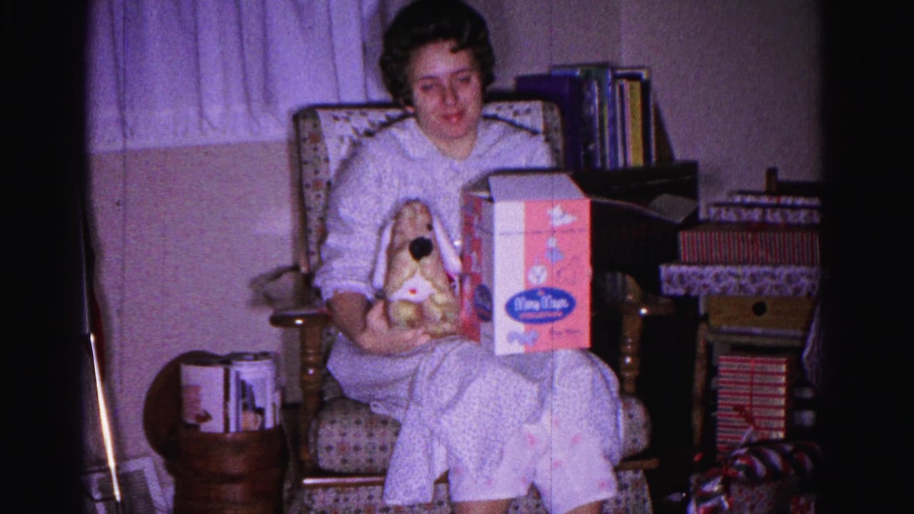 8mm home-movie frame, Hagerstown Maryland c.1962: woman in pajamas on Christmas morning holding a stuffed rabbit and a branded toy box in a domestic living room.