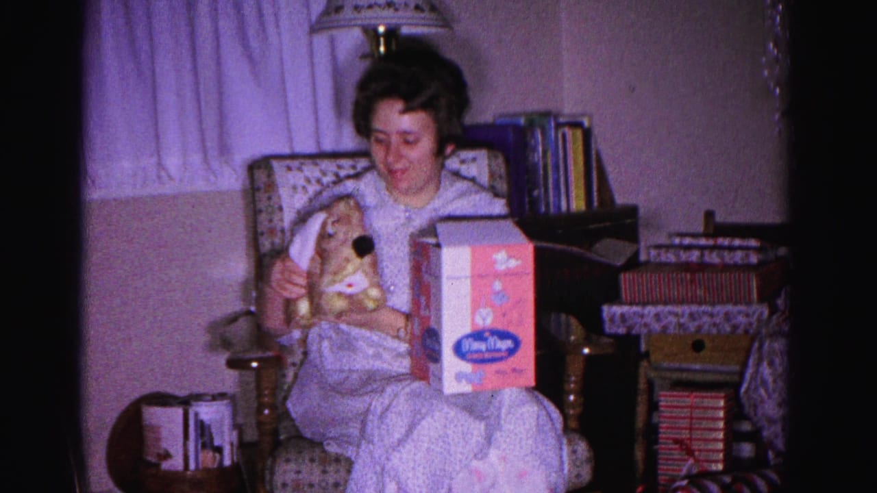 8mm frame c.1962: woman in pajamas displaying Christmas gifts — a stuffed rabbit and branded toy box — in a domestic Maryland interior, books and curtains behind her.