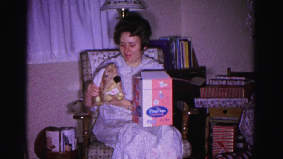 8mm frame c.1962: woman in pajamas displaying Christmas gifts — a stuffed rabbit and branded toy box — in a domestic Maryland interior, books and curtains behind her.