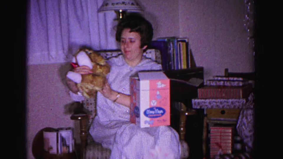 8mm frame c.1962: woman in pajamas examining a stuffed animal and branded toy box in a domestic interior on Christmas morning, bookshelf visible behind her.