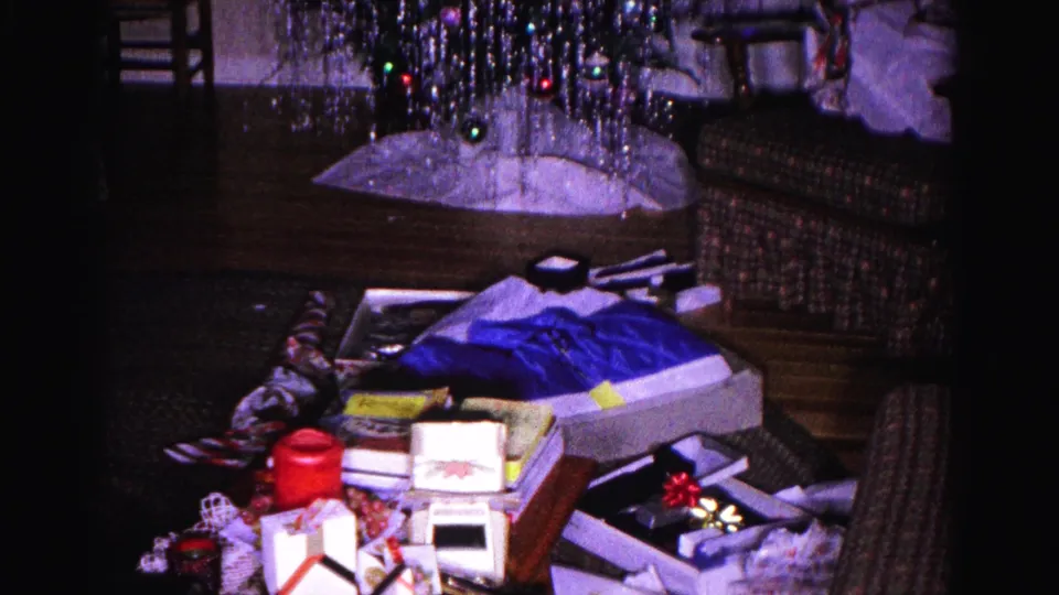 8mm wide shot c.1962: Christmas tree hung with silver tinsel and colored string lights, surrounded by scattered opened gifts and boxes on a domestic living-room floor.