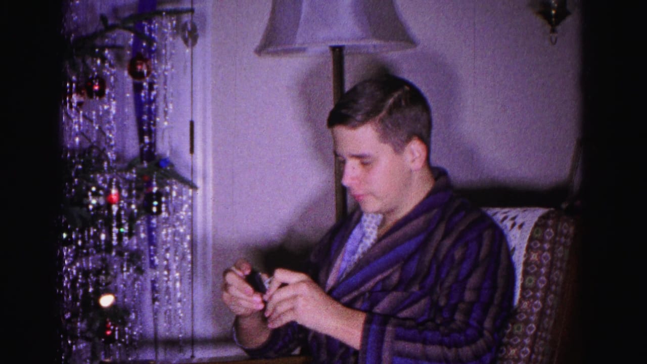 Young man in dark striped bathrobe examines a small gift, 8mm home movie Christmas morning 1962, Hagerstown Maryland; silver tinsel tree and wall lamp visible behind him.
