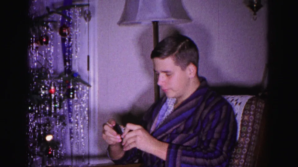 Young man in dark striped bathrobe examines a small gift, 8mm home movie Christmas morning 1962, Hagerstown Maryland; silver tinsel tree and wall lamp visible behind him.