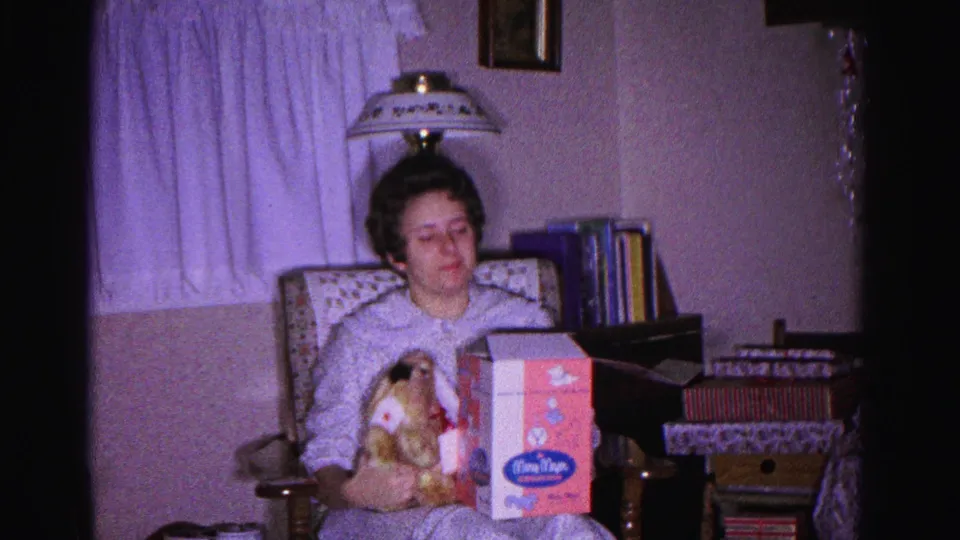 Woman in light nightgown holds a stuffed animal and a pink branded gift box on Christmas morning; table lamp, bookshelves, and wrapped presents visible behind her; 8mm home movie, Hagerstown Maryland, 1962.