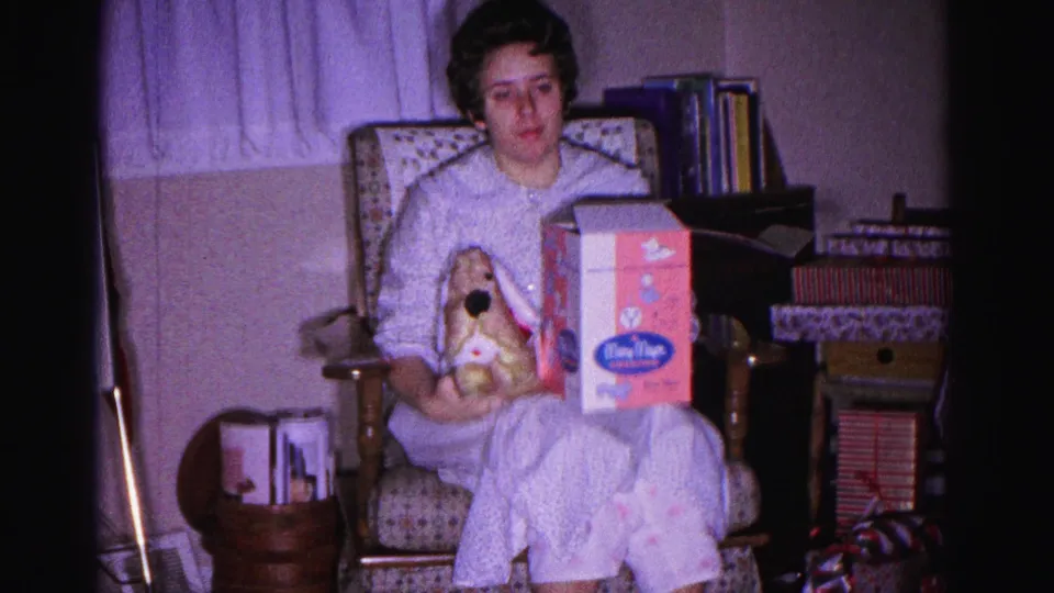 Woman in light pajamas displays a stuffed rabbit and a pink cosmetics box for the camera; bookshelves and wrapped presents visible in background; 8mm home movie, Christmas 1962, Hagerstown Maryland.