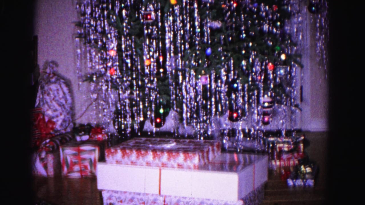 Aluminum tinsel Christmas tree with colored lights and ribbon-wrapped gifts beneath, in a 1960s living room interior.