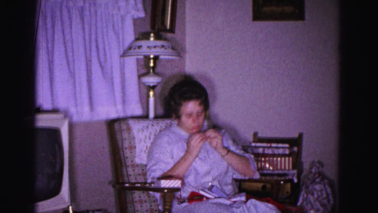 Woman in light housecoat doing needlework in a patterned armchair, table lamp with decorative shade and sheer curtains behind, circa 1962 home interior, Hagerstown, Maryland.