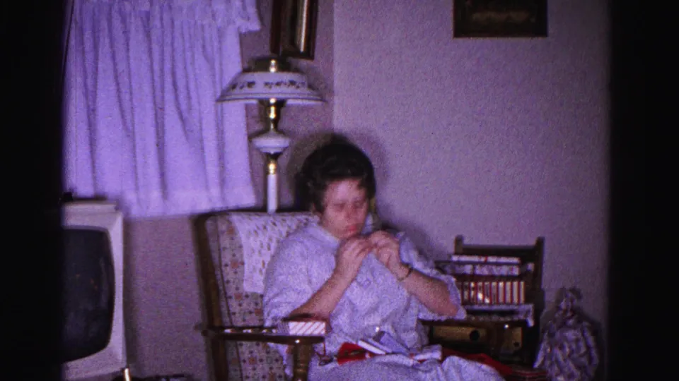 Woman in light housecoat doing needlework in a patterned armchair, table lamp with decorative shade and sheer curtains behind, circa 1962 home interior, Hagerstown, Maryland.