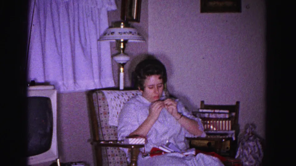Wider view of woman knitting in a patterned armchair in a 1960s living room, vintage television at left edge, table lamp, secondary chair, and wrapped gifts partially visible at right, circa 1962.