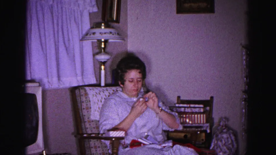 Woman absorbed in knitting or crocheting in a patterned armchair, table lamp with decorative shade and sheer curtains visible behind, circa 1962 domestic interior, Hagerstown, Maryland.