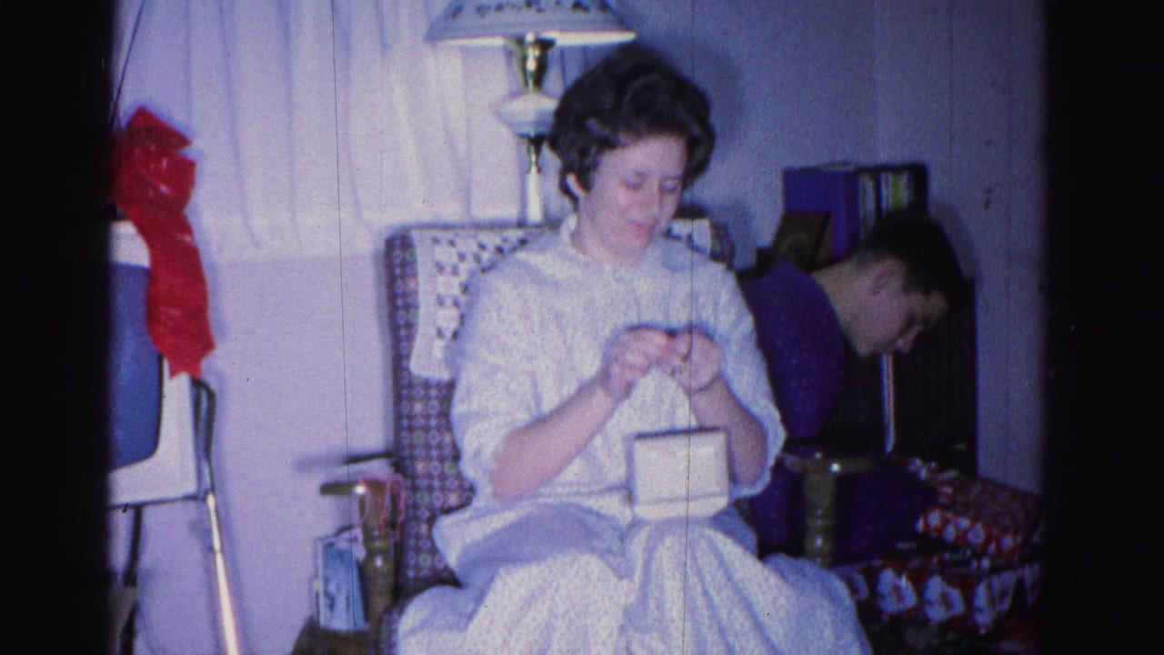 Woman in white dress examining a small white box while seated in a wicker armchair; a second person is visible behind her to the left.