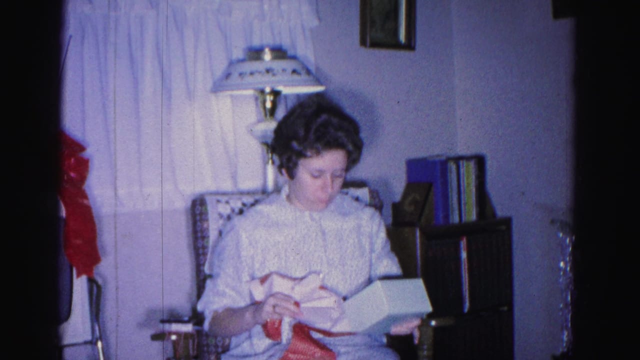 Woman seated in wicker armchair holding an open white gift box with red ribbon or wrapping; a large red bow is visible to the left.