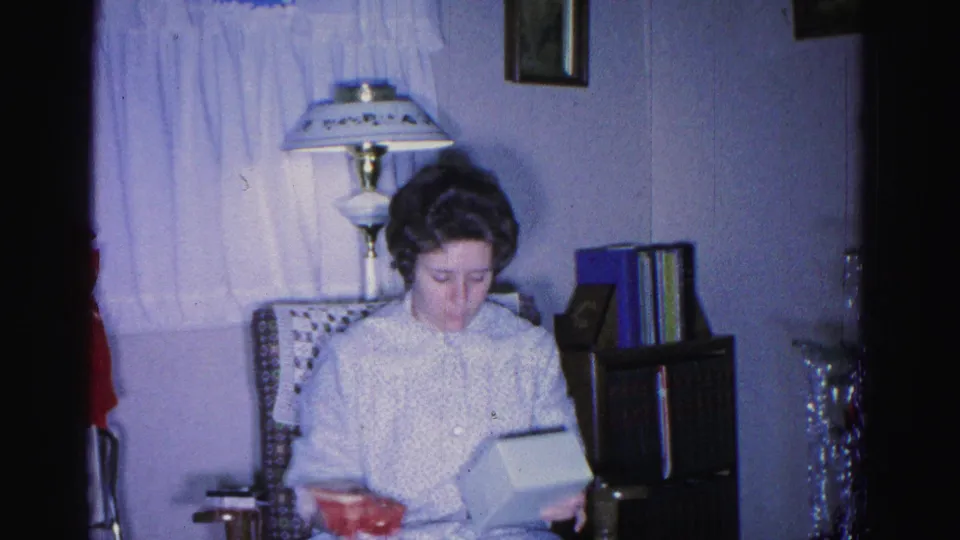 1962 home movie, Hagerstown Maryland — woman examining the contents of an opened gift box in a mid-century living room with silver tinsel and red bow visible at frame edges.