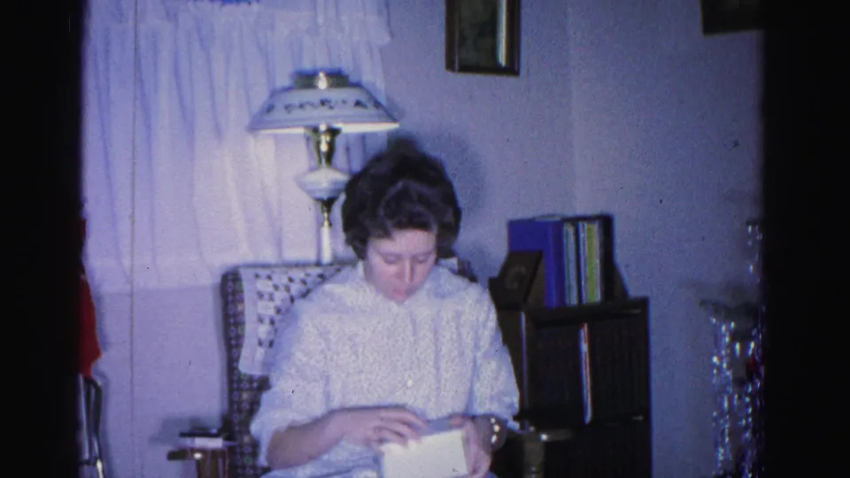1962 home movie, Hagerstown Maryland — woman in white patterned dress continues examining gift box in a mid-century domestic interior with holiday decorations at frame edges.