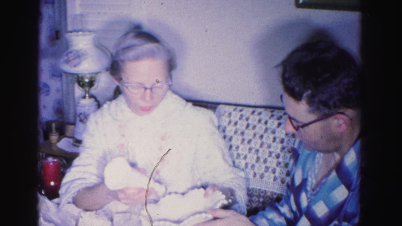 Elderly woman with glasses cradles a bundled infant while a man in a blue checkered shirt leans in, domestic interior, 1962 home movie, Hagerstown, Maryland.