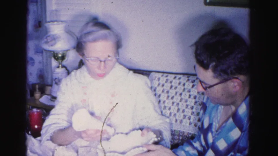 Elderly woman with glasses cradles a bundled infant while a man in a blue checkered shirt leans in, domestic interior, 1962 home movie, Hagerstown, Maryland.