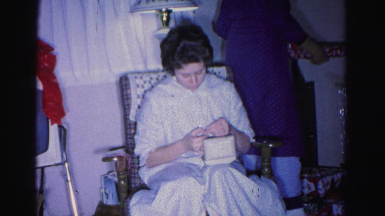 Younger woman with dark hair in a white patterned dress seated in a patterned wingback chair, working on handiwork; lamp and red decorative items visible in background.