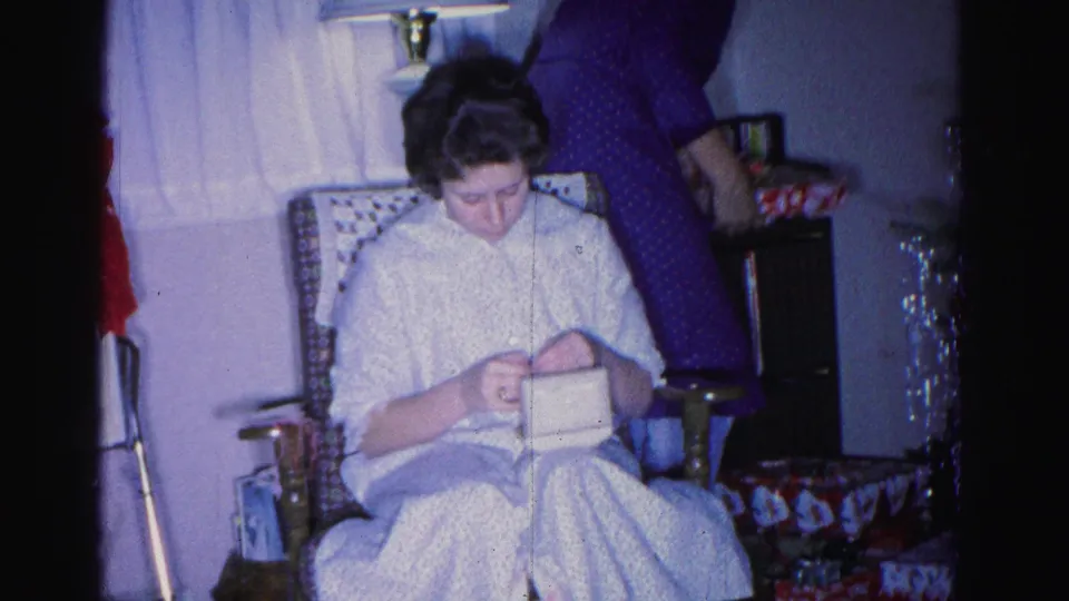 Younger woman knitting or sewing in a patterned wingback chair; man in blue checkered shirt and red decorative items visible in background, 1962 home movie.