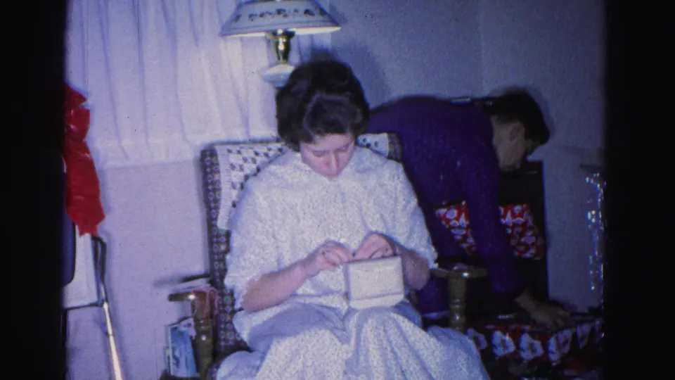 Younger woman continues handiwork in a patterned wingback chair; man at right edge and red decorations visible behind her, 1962 home movie interior.