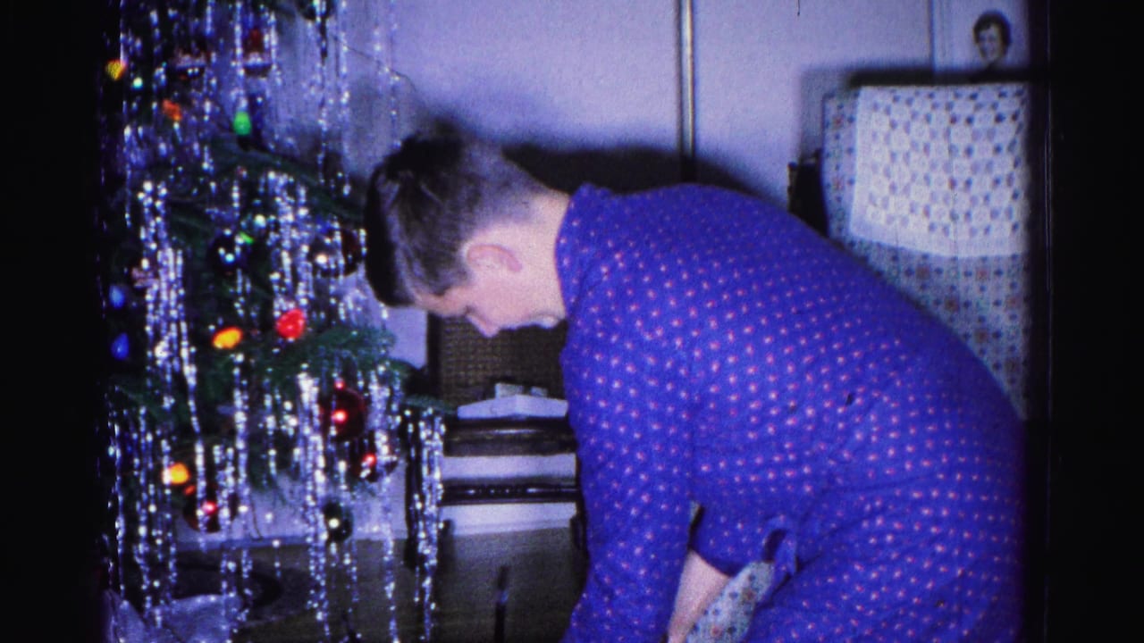 Young man in blue polka-dot pajamas bending over gifts beneath a tinsel-decorated Christmas tree in a living room.
