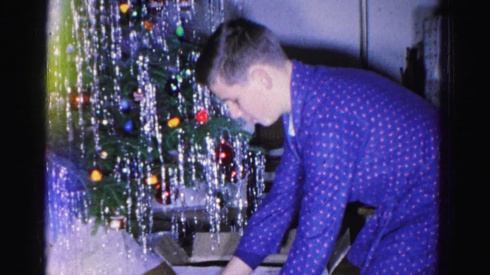 Young man crouching beside a heavily tinsel-draped Christmas tree, handling gifts at floor level, mid-century American home interior, circa 1962.