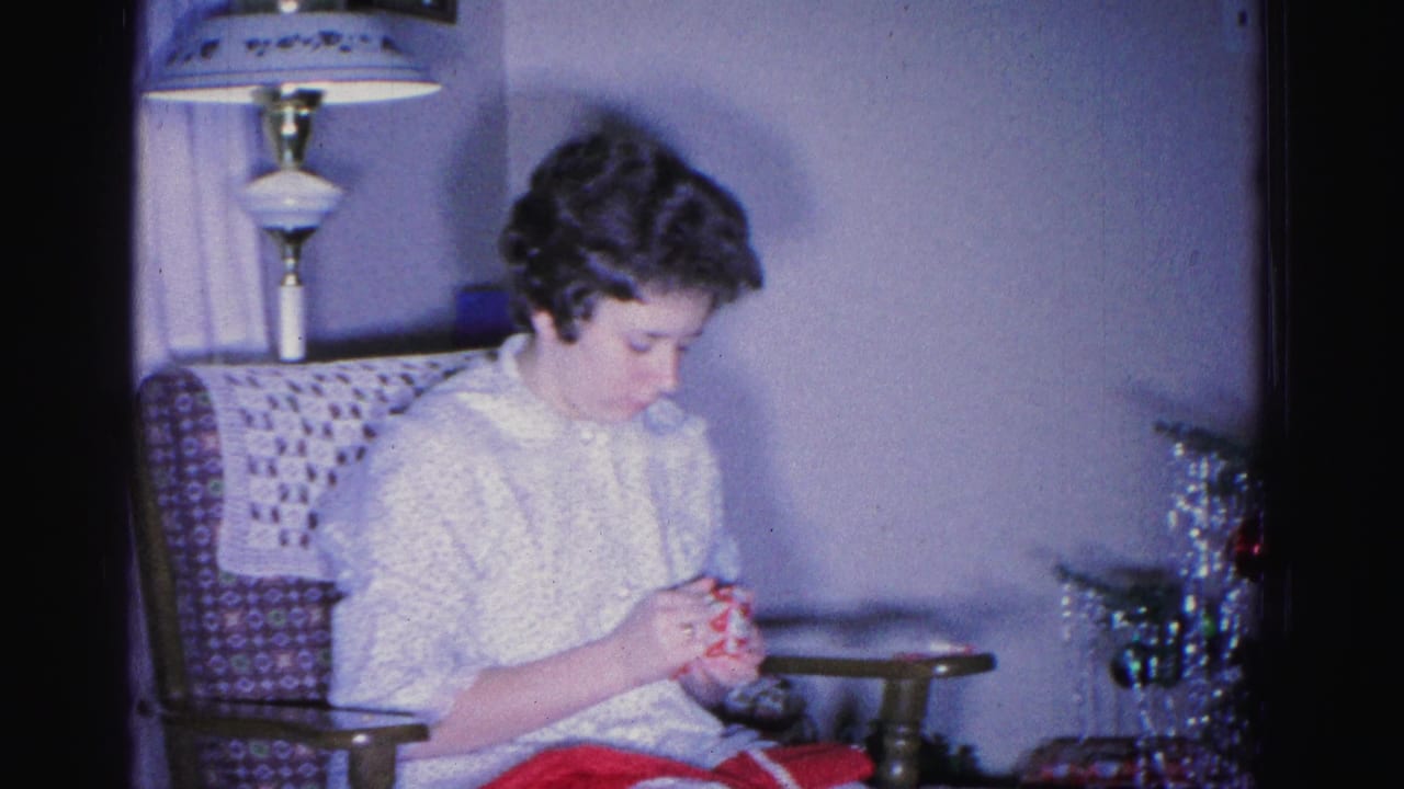 Woman crocheting red yarn in a patterned armchair, tinsel-decorated Christmas tree and table lamp visible behind her; 8mm home movie, circa 1962, Hagerstown, Maryland.