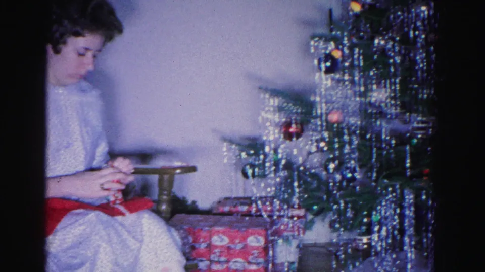 Woman in white top crocheting at left; silver-tinsel Christmas tree with colored ornaments and gift-wrapped packages at base dominate right; 8mm home movie, circa 1962, Hagerstown, Maryland.
