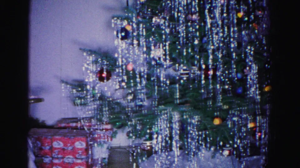 Tinsel-laden Christmas tree with colorful ornaments and wrapped gift packages at base, crocheting woman at left edge; 8mm home movie, circa 1962, Hagerstown, Maryland.