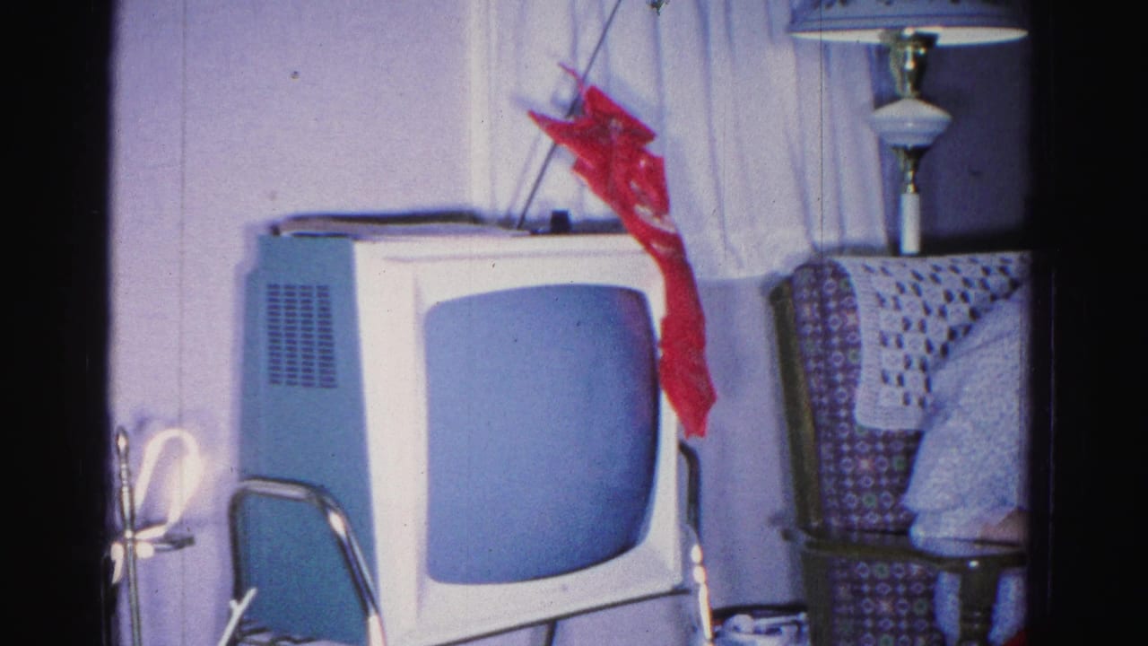 Vintage boxy television set with a blank blue-grey screen and rabbit-ear antenna; red ribbon bow on top, patterned armchair and floor lamp visible at right.