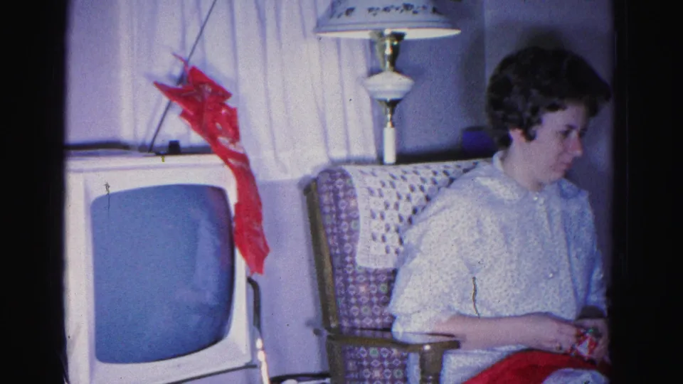 A young woman seated in an upholstered armchair examines an object (possibly a gift) in a Christmas-decorated 1960s living room, Hagerstown, Maryland, circa 1962.