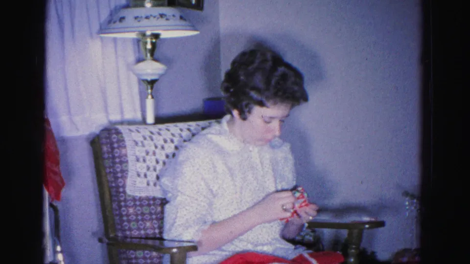 A young woman examines a small object (possibly a gift) while seated in a patterned armchair in a 1960s living room; floor lamp and curtains visible, Hagerstown, Maryland, circa 1962.
